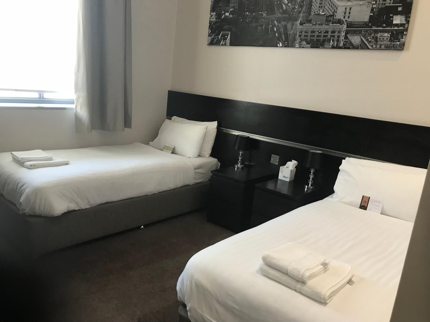 A hotel room with two beds, a nightstand with a lamp, and a cityscape painting on the wall.