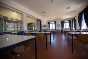 A cafeteria with multiple tables and chairs, a counter with a display case, and a window with curtains. - HI Oeiras - Pousada de Juventude - Hostel (Oeiras)