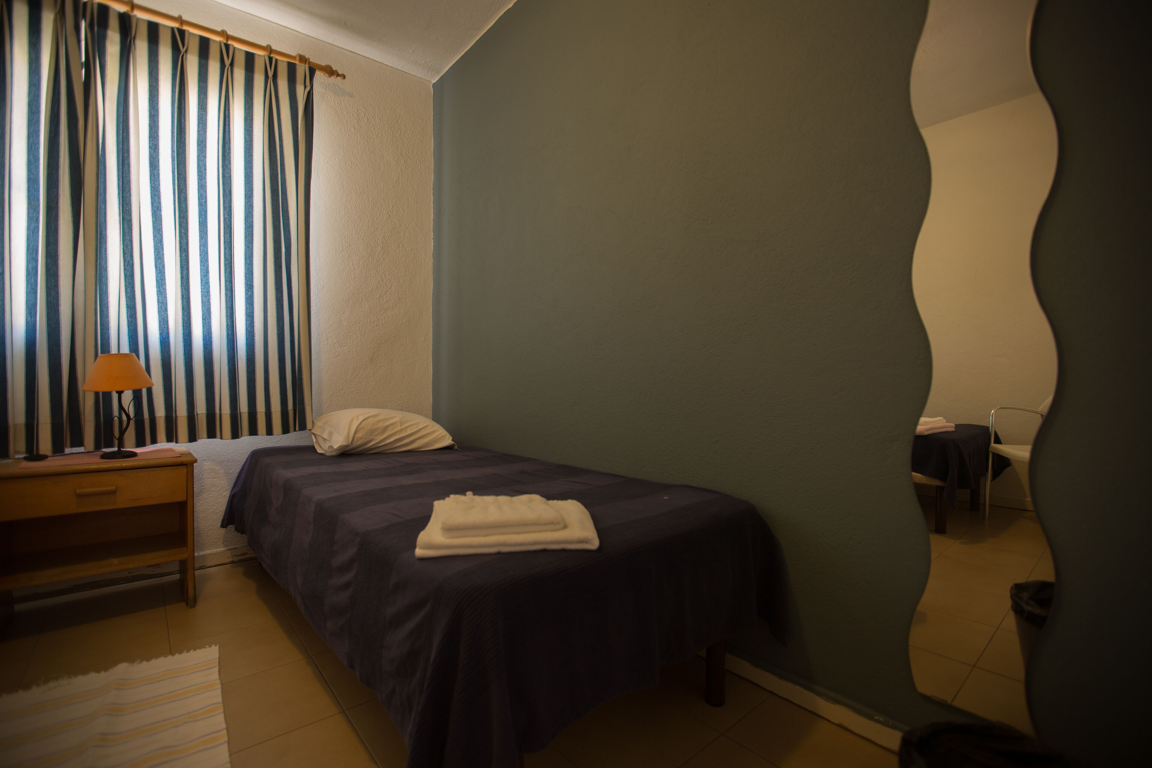 A bedroom with a bed, a nightstand, a lamp, a mirror, and a window with curtains.