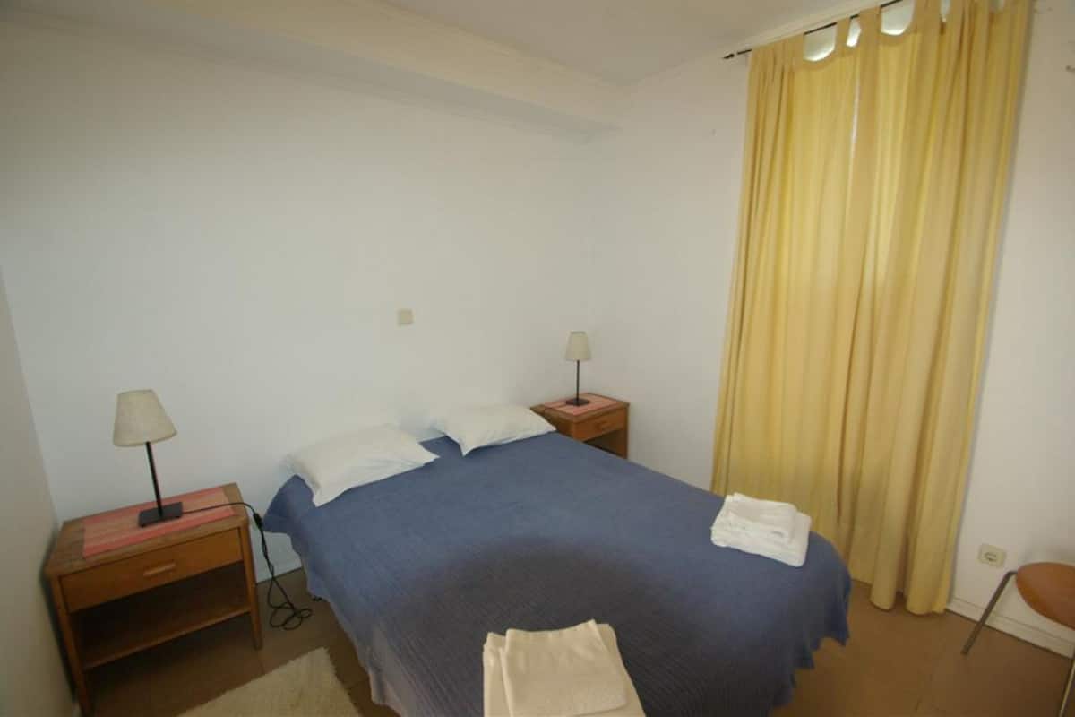 A bedroom with a bed, two bedside tables, a lamp on each table, a window with yellow curtains, and a chair.