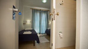 A hotel room with two beds, a bathroom with a toilet paper holder, and a striped curtain. - HI Oeiras - Pousada de Juventude - Hostel (Oeiras)