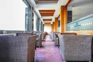 A row of wicker chairs with a wooden floor and large windows along a corridor. - Hotel Subash International (Reasi)