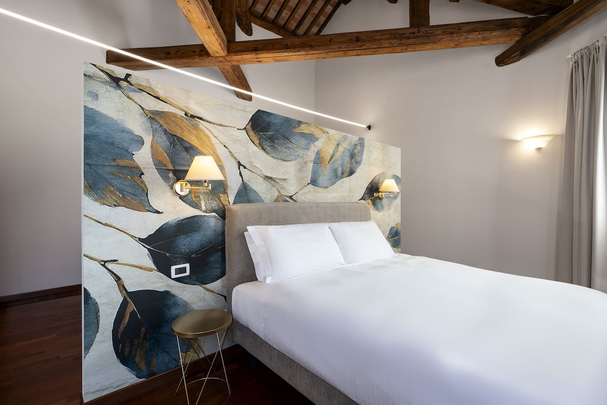 A modern bedroom with a large bed, a headboard with a leaf pattern, two wall-mounted lamps, and a wooden ceiling beam.