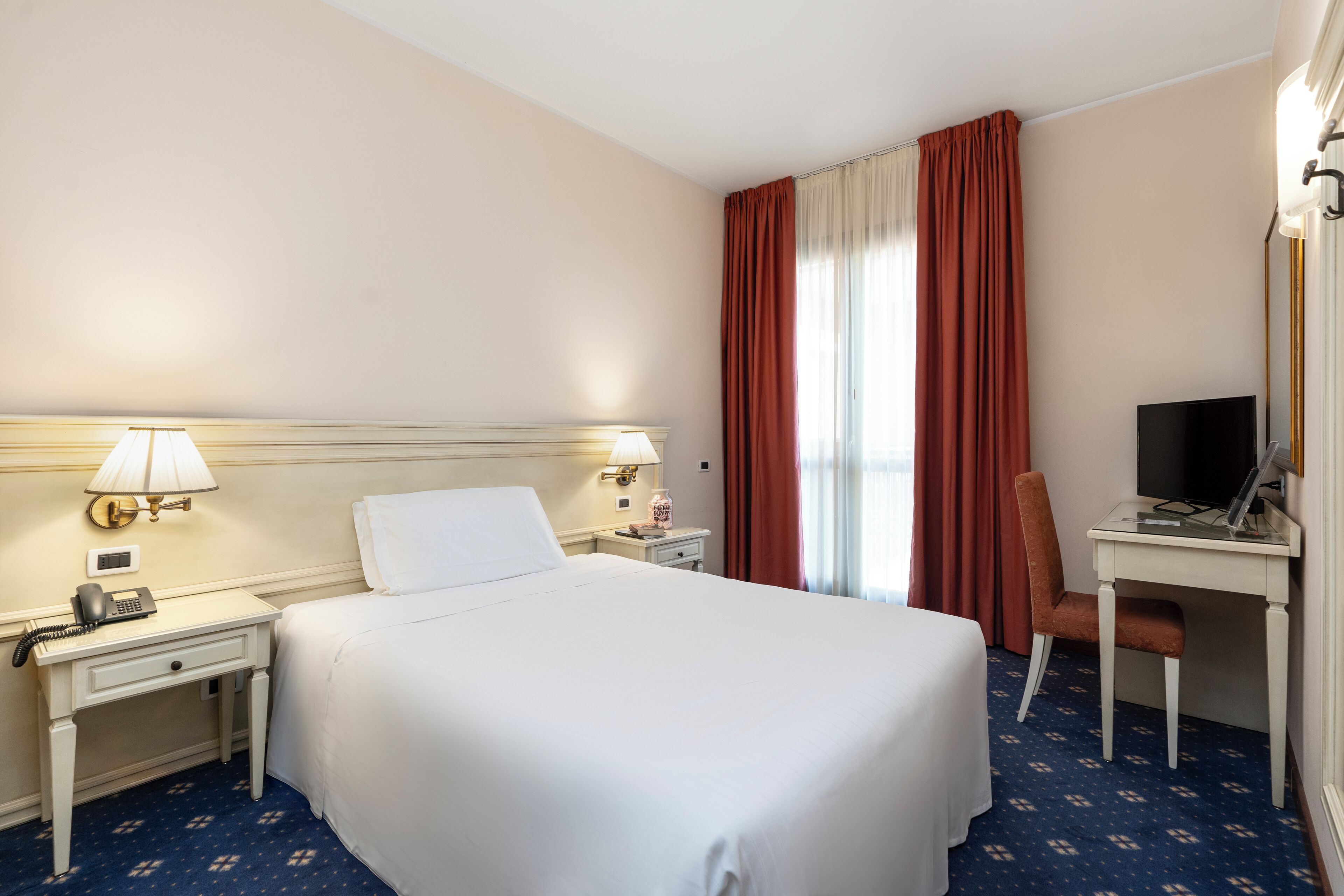 Hotelkamer met een bed, een bureau met een computer, een stoel, een lamp, een telefoon en een raam met gordijnen.