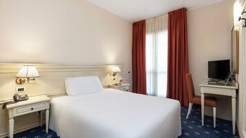 Hotelkamer met een bed, een bureau met een computer, een stoel, een lamp, een telefoon en een raam met gordijnen.
