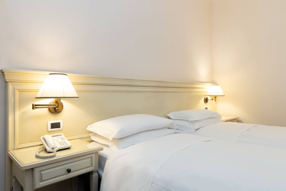 A hotel room with a bed, two bedside lamps, a nightstand with a telephone, and a wall-mounted light switch.