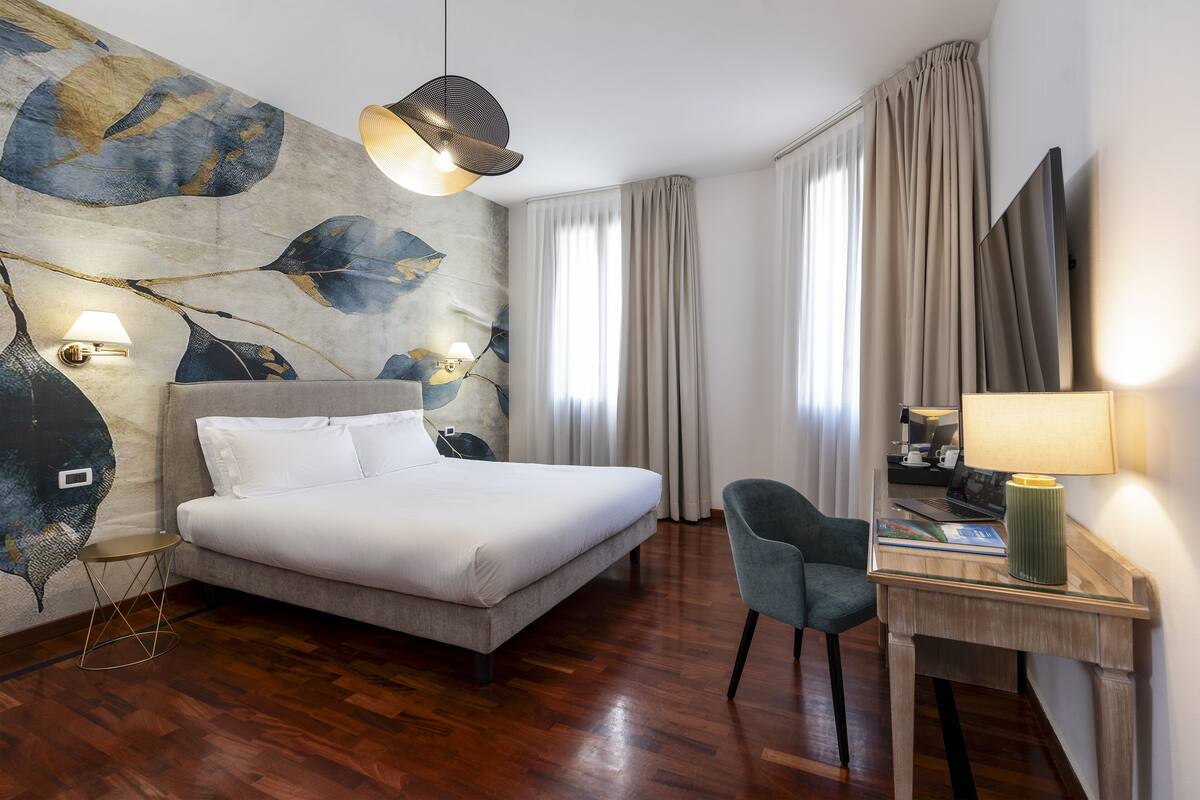 A hotel room with a large bed, a desk with a lamp, a chair, and a wall with a leaf pattern.