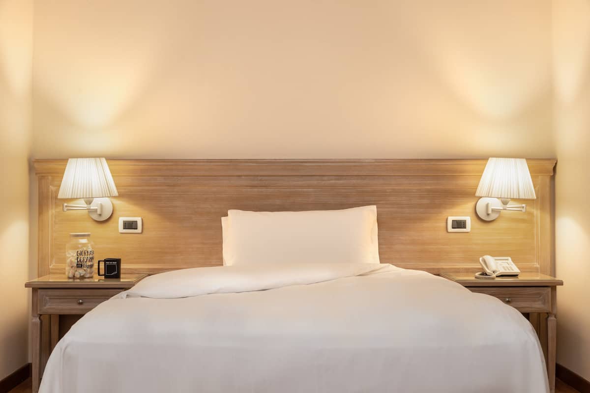 A neatly made bed with white linens, two bedside lamps, a nightstand with a phone, a small table with a jar of cookies, and a thermostat on the wall.