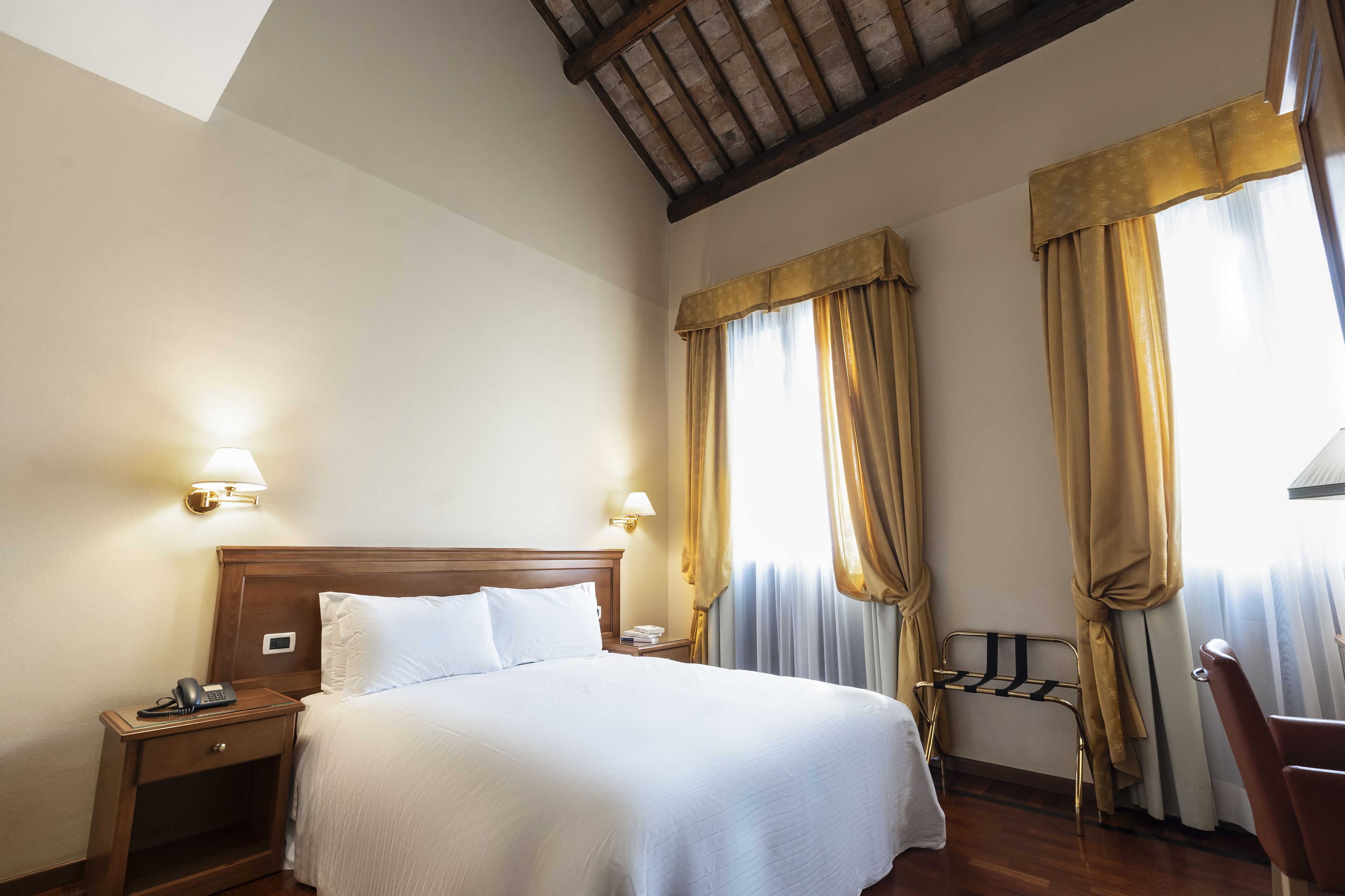 Una habitación de hotel con cabecera de madera, una cama individual, dos lámparas de noche, una mesita de noche con un teléfono y una lámpara, y una ventana con cortinas.
