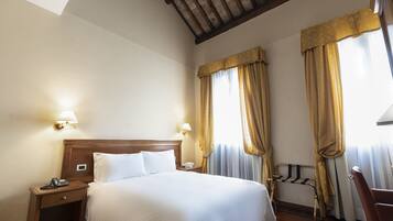 Una habitación de hotel con cabecera de madera, una cama individual, dos lámparas de noche, una mesita de noche con un teléfono y una lámpara, y una ventana con cortinas.