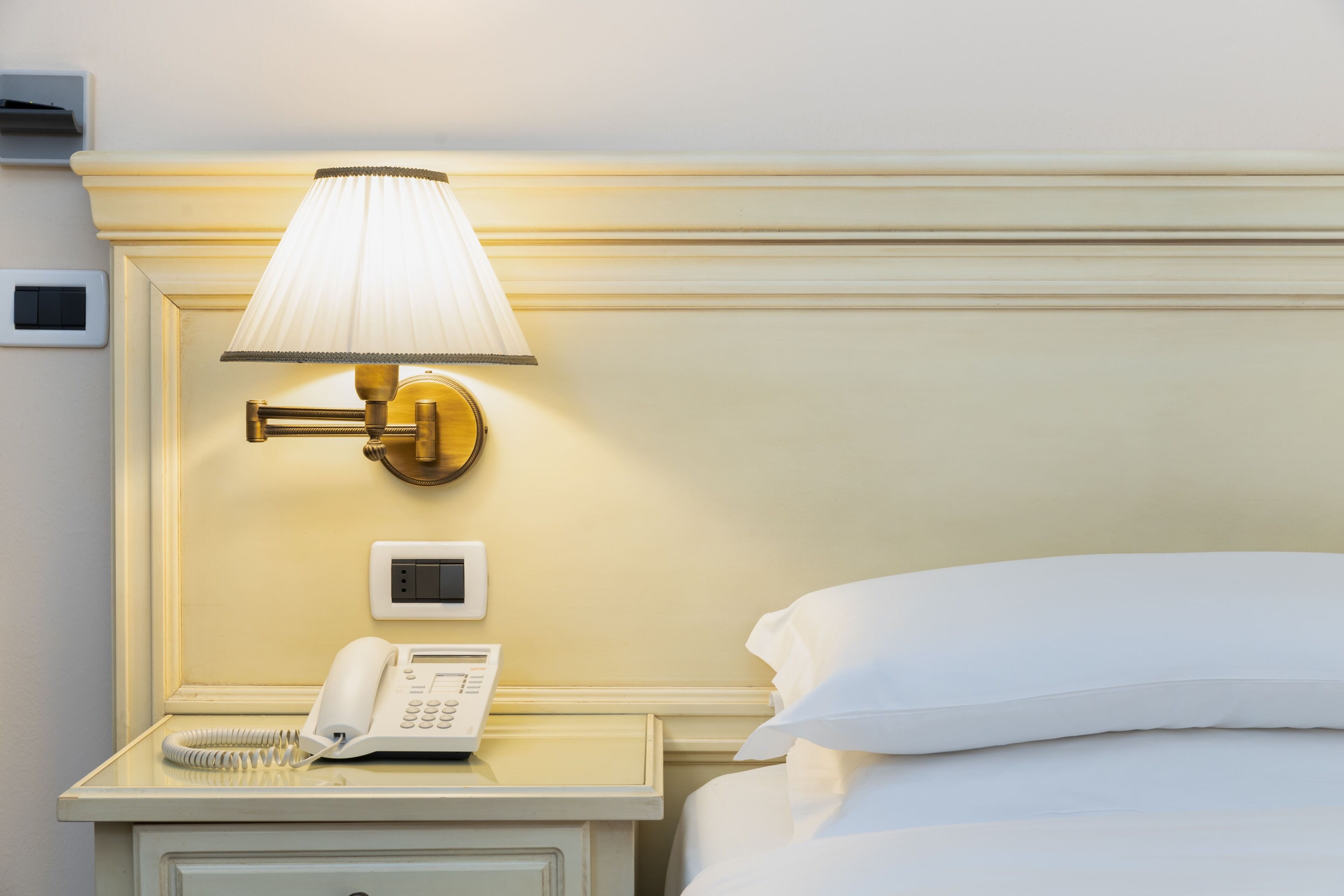 A hotel room with a bedside table, a lamp, a phone, and a bed with white bedding.