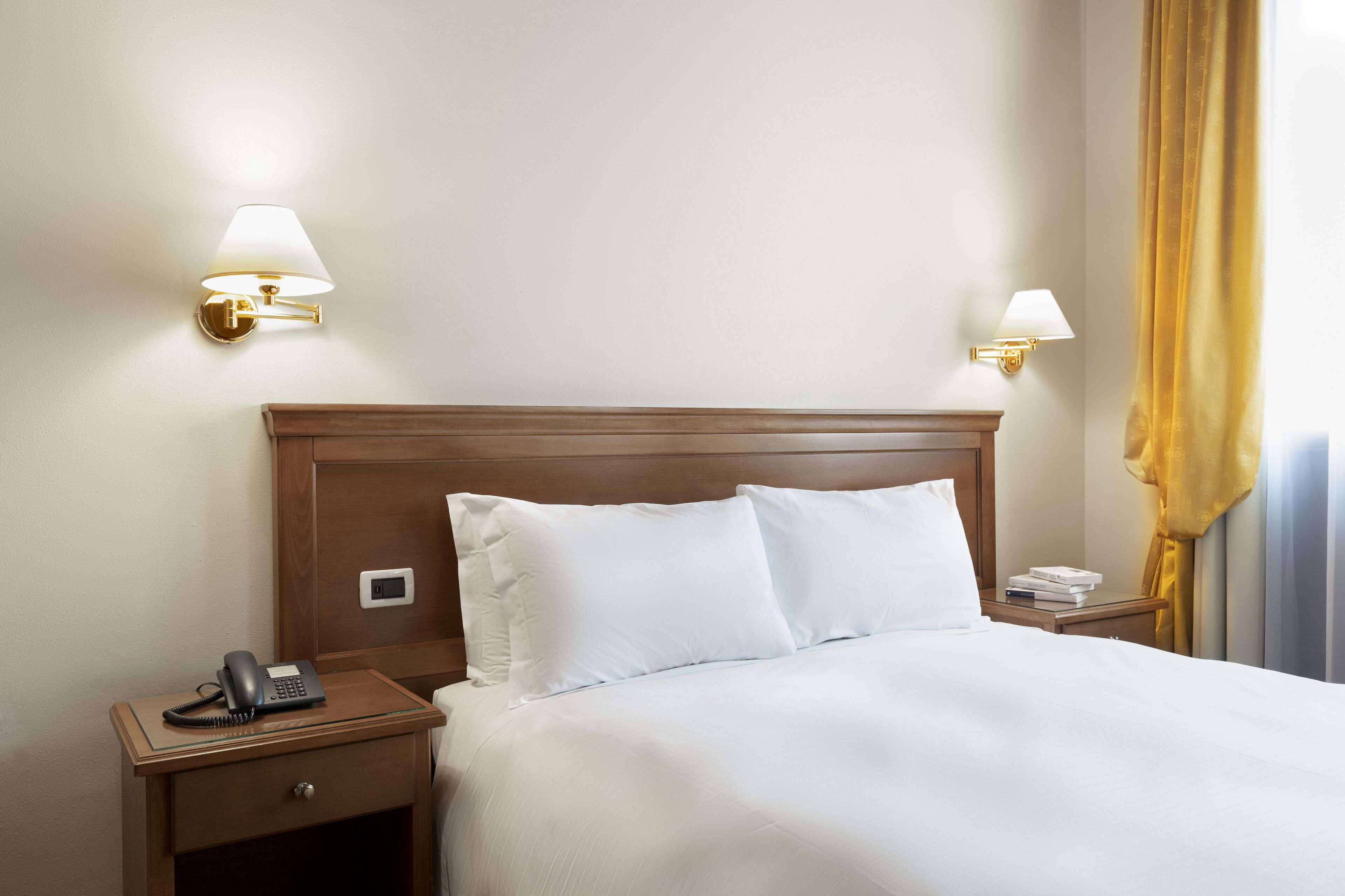 Een hotelkamer met een houten hoofdeinde, twee bedlampen, een telefoon en een nachtkastje met een boek en een afstandsbediening.