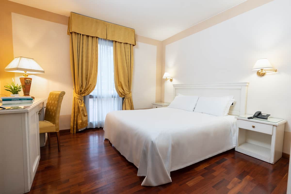A hotel room with a bed, bedside table, lamp, chair, and a desk with a plant.