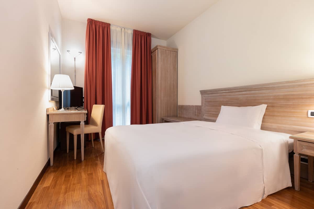 A hotel room with a bed, a desk with a chair, a lamp, and a wardrobe.