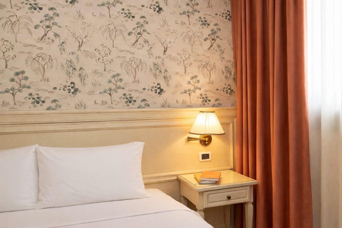 A hotel room with a bed, a nightstand with a lamp, and a wall with floral wallpaper.