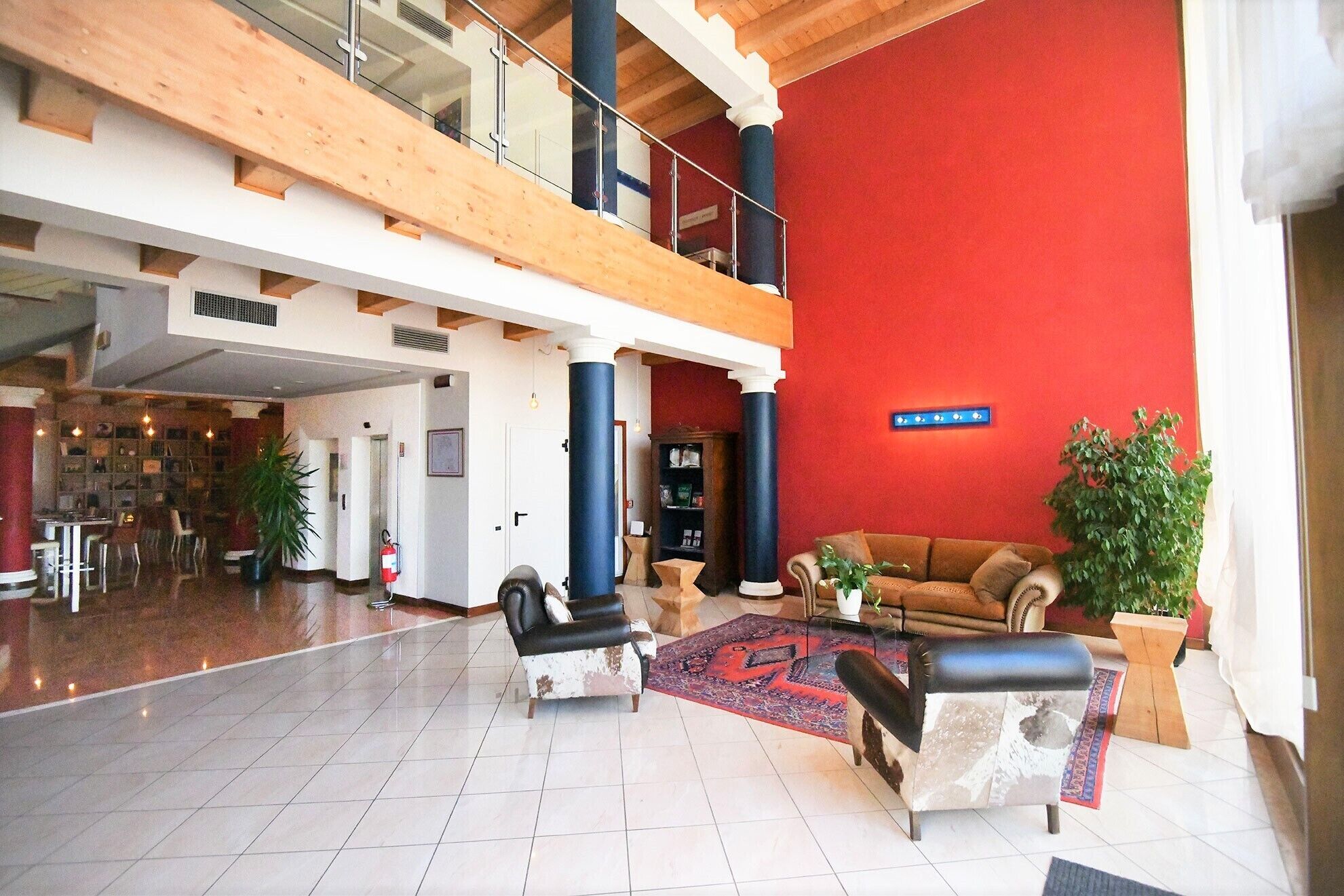 A spacious lobby with a high ceiling, a red accent wall, a seating area with a sofa and armchairs, and a staircase leading to an upper level.
