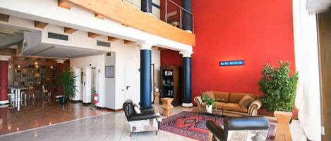 A spacious lobby with a high ceiling, a red accent wall, a seating area with a sofa and armchairs, and a staircase leading to an upper level.