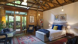 Une chambre avec un grand lit, une chaise, une table et une bibliothèque. On peut admirer la vue sur l’extérieur à travers une porte vitrée.