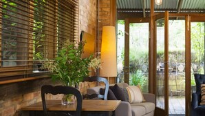 Un espace intérieur accueillant avec une table en bois, une chaise et un canapé. Il y a une fenêtre avec des stores, une lampe et une vue sur l’extérieur.