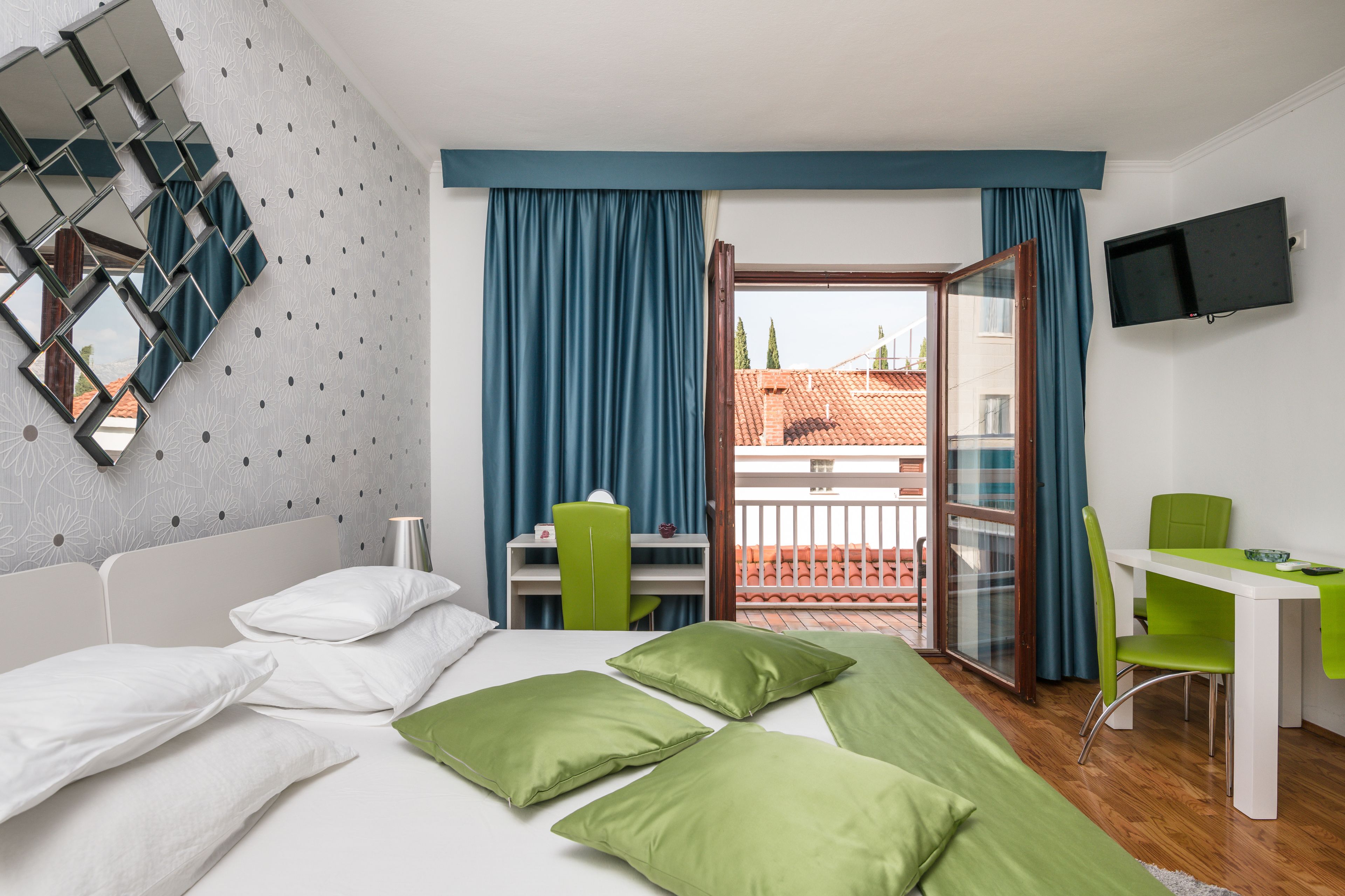 Habitación de hotel con cama, almohadas verdes, escritorio, silla, televisión y balcón con cortinas azules.