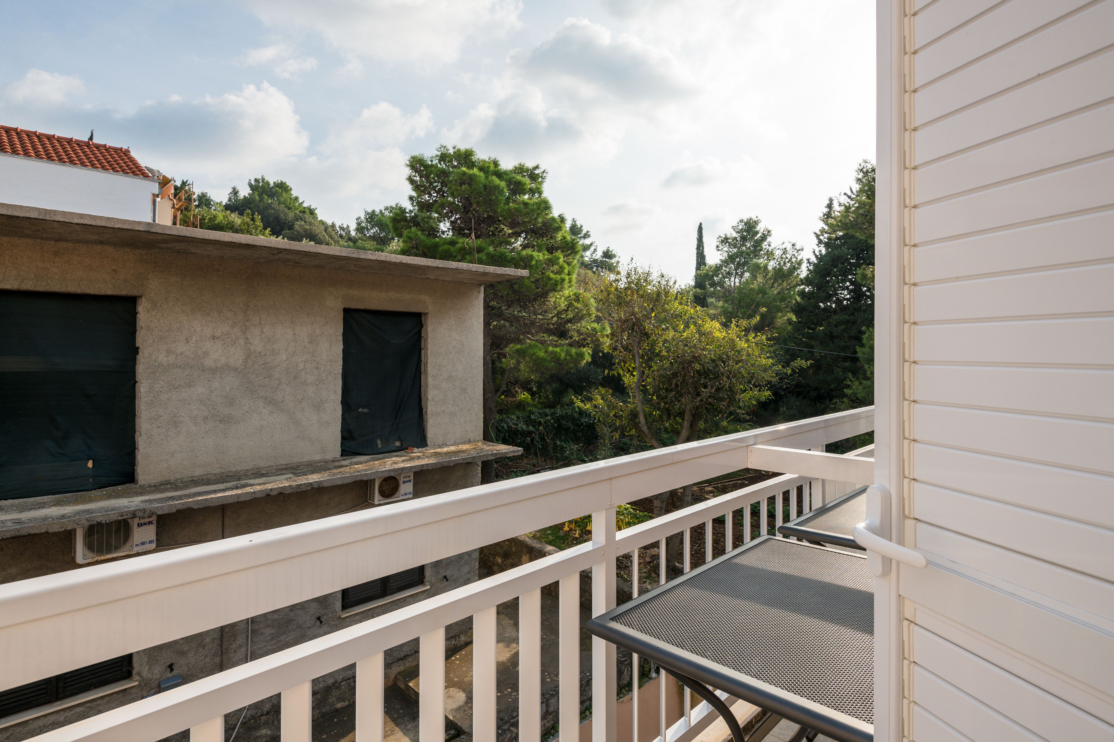 A balcony with a white railing, a concrete structure with a black door, and a view of trees and a building with a red roof.