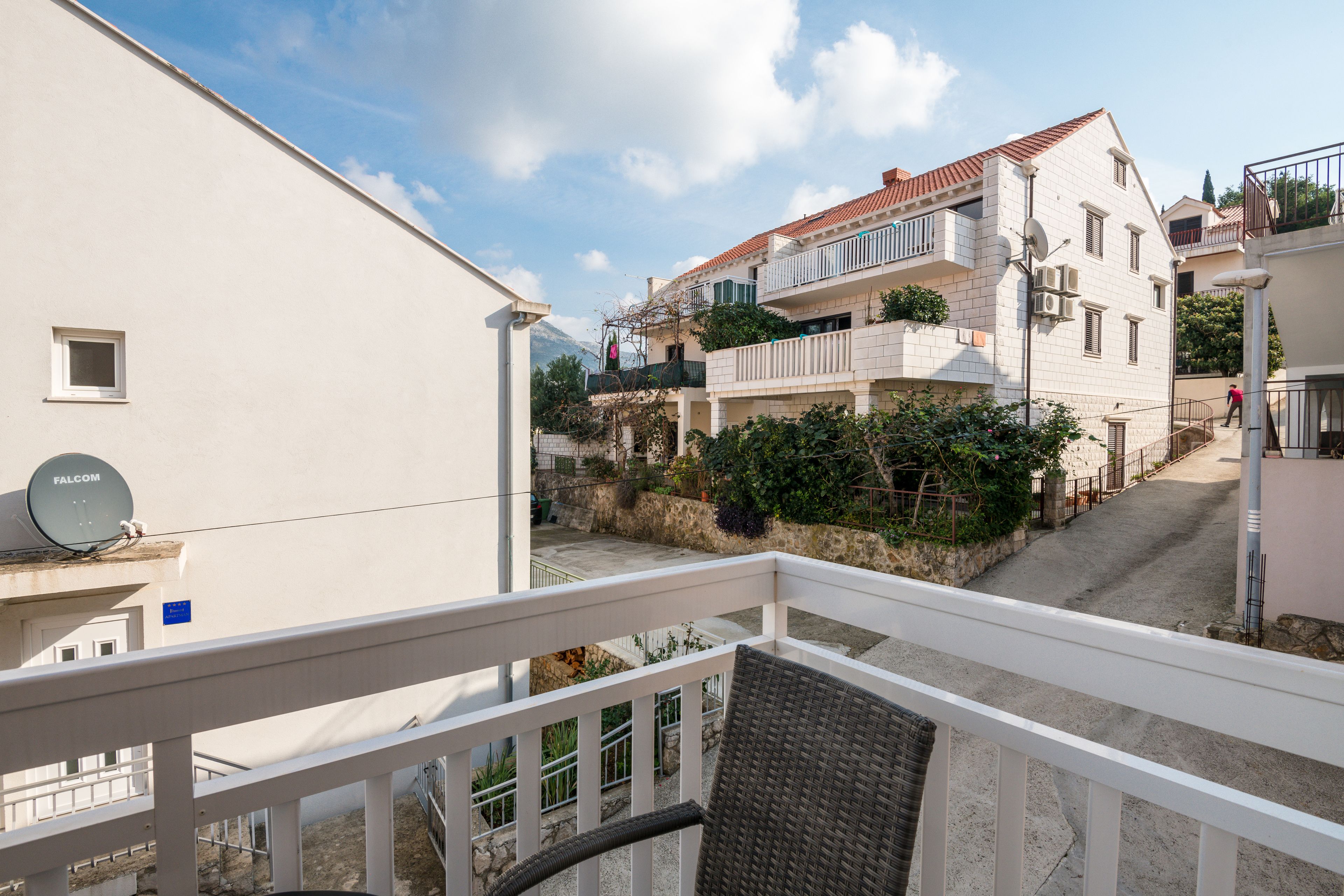 A balcony with a wicker chair, a white railing, and a view of a residential street with buildings and greenery.