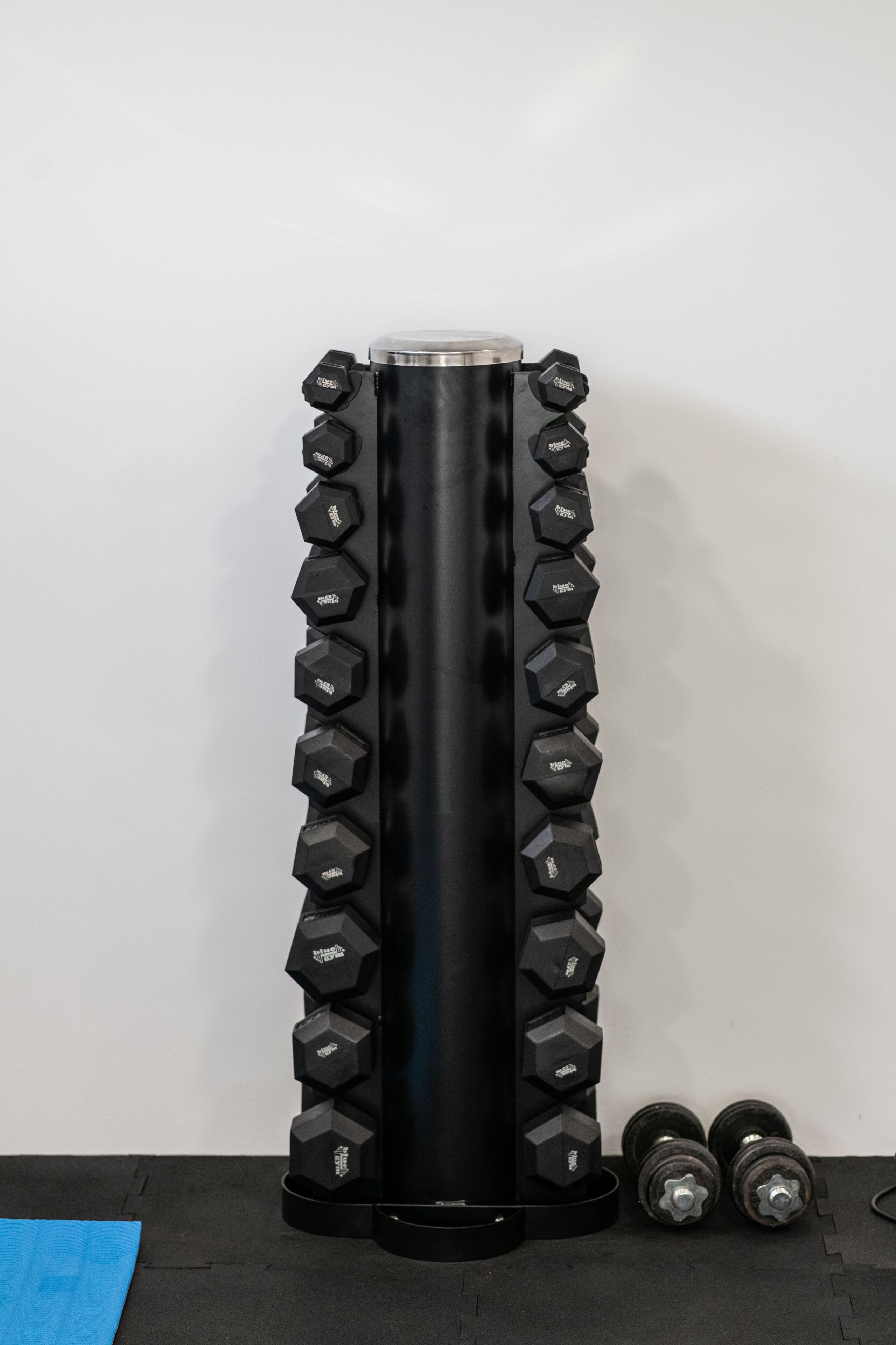 Un soporte para mancuernas negro con agarres hexagonales y una parte superior metálica, dos mancuernas sobre el suelo a su lado, y una colchoneta azul al fondo.