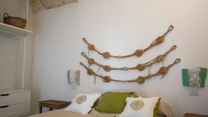 Un dormitorio con cama, almohadas, una mesita de madera, una mesita de noche, una obra de arte rústica en la pared y un estante con una cesta.