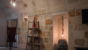 Una habitación rústica con paredes de piedra, un estante de madera con escalera, una lámpara colgante y un jarrón grande.