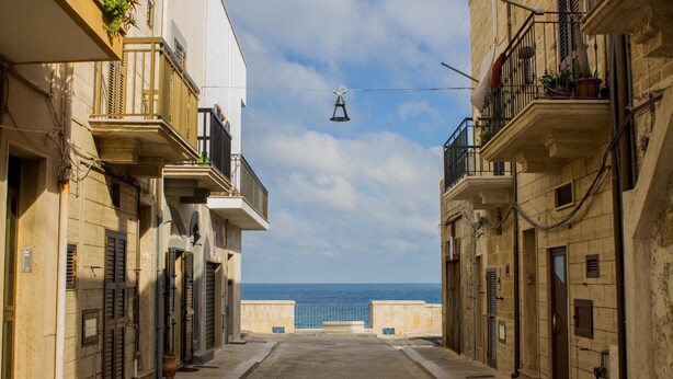 Una calle estrecha flanqueada por edificios, cielo despejado y vista al mar en la distancia.