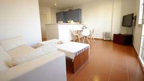 A living room with a sofa, a dining table with chairs, a kitchen area with cabinets, and a television.