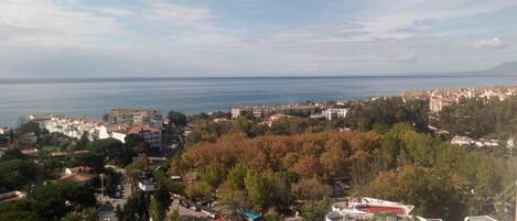 A coastal town with a view of the sea. There are residential buildings, a park with palm trees, and a multi-story building with balconies.