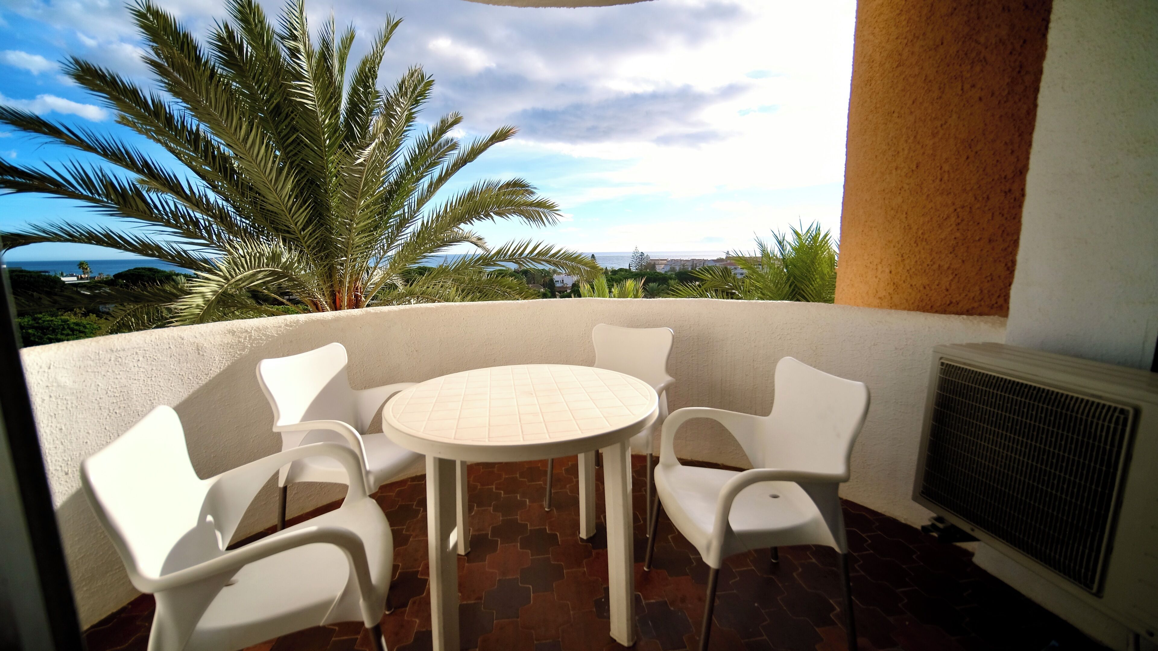 Un balcon avec une table ronde et quatre chaises, un radiateur et une vue sur l’océan et les palmiers.