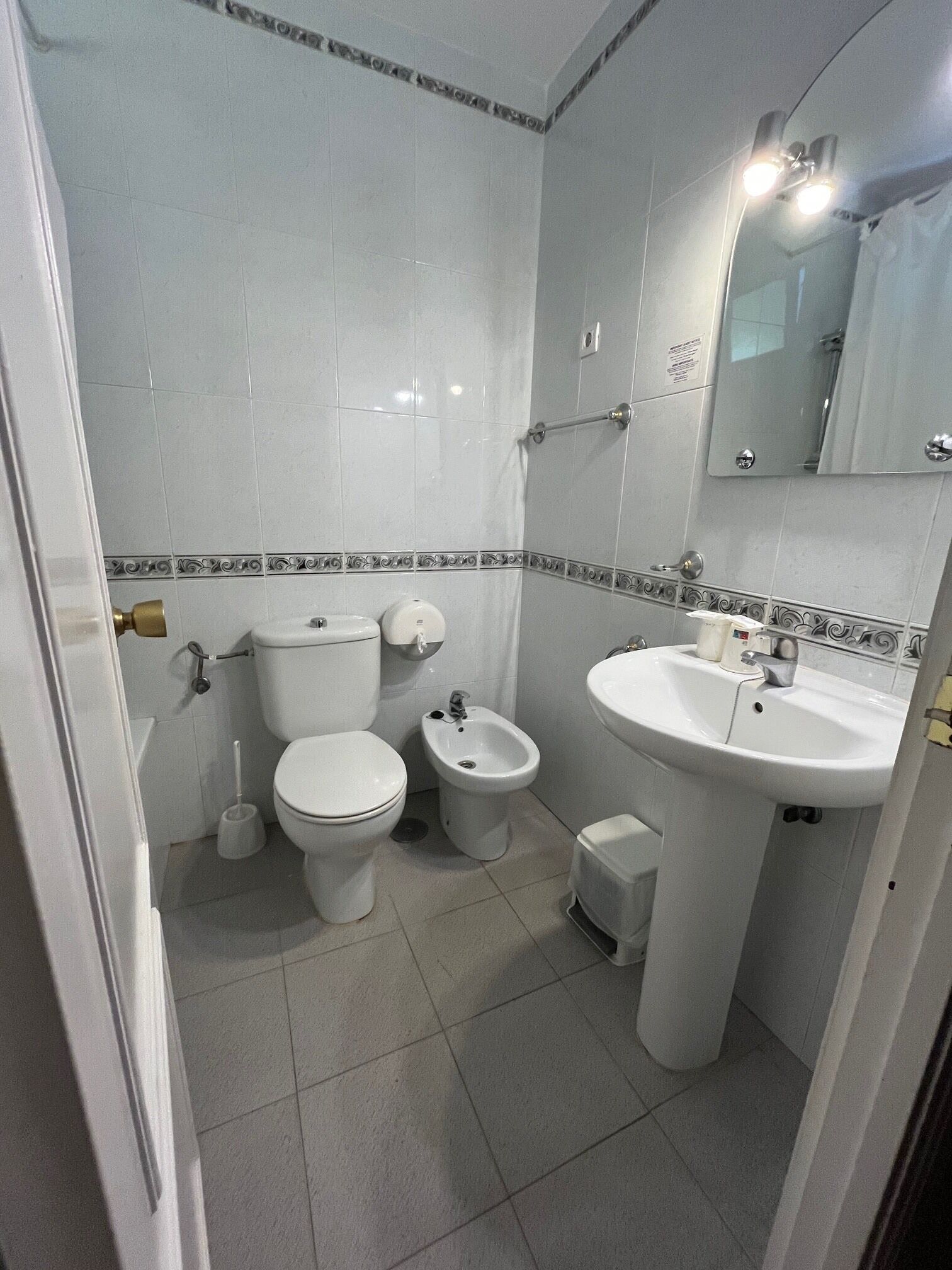 A bathroom with a toilet, bidet, and sink, all in white, with a tiled floor and a mirror above the sink.