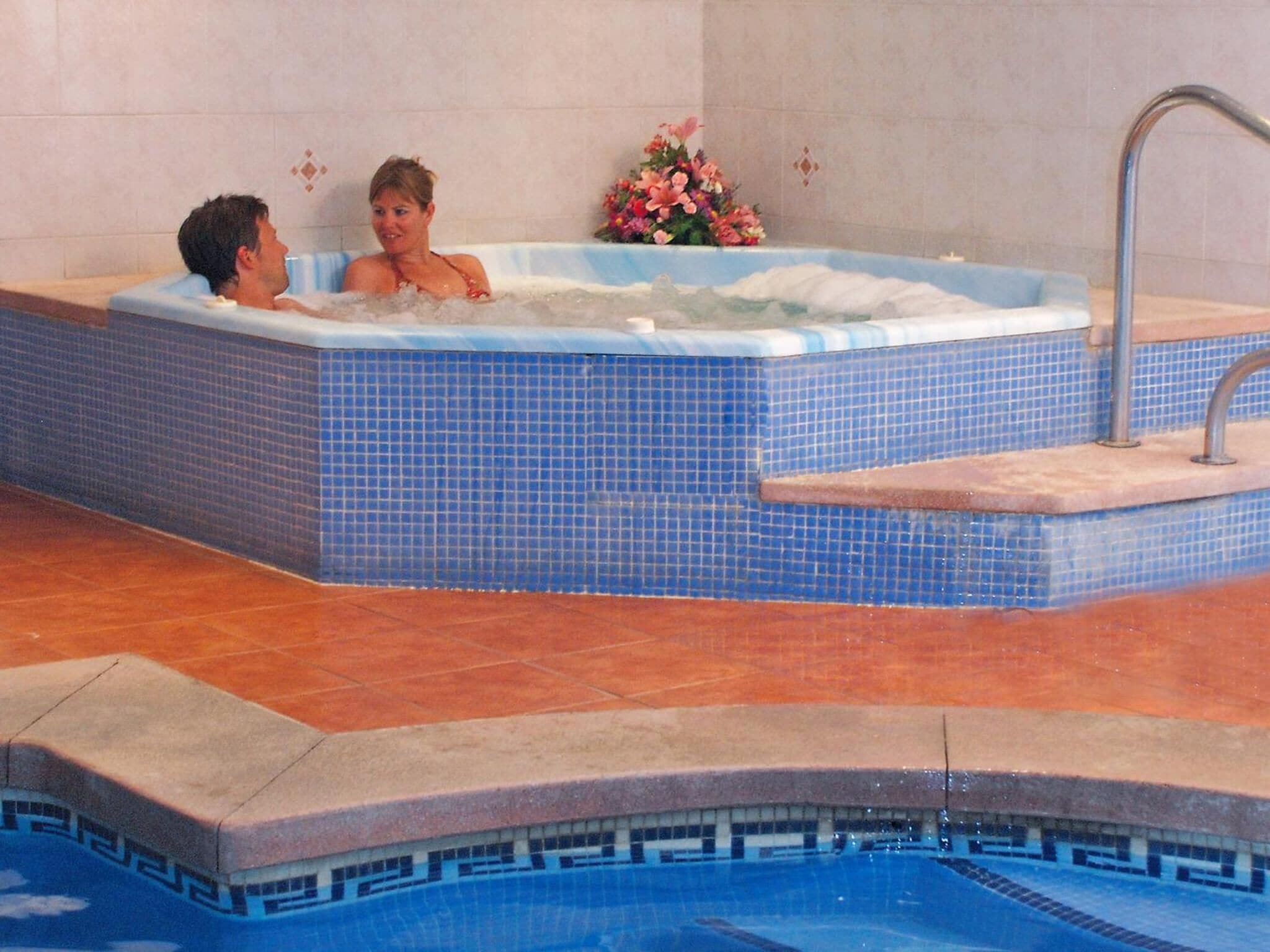 A hot tub with two people, a tiled wall, and a flower arrangement.