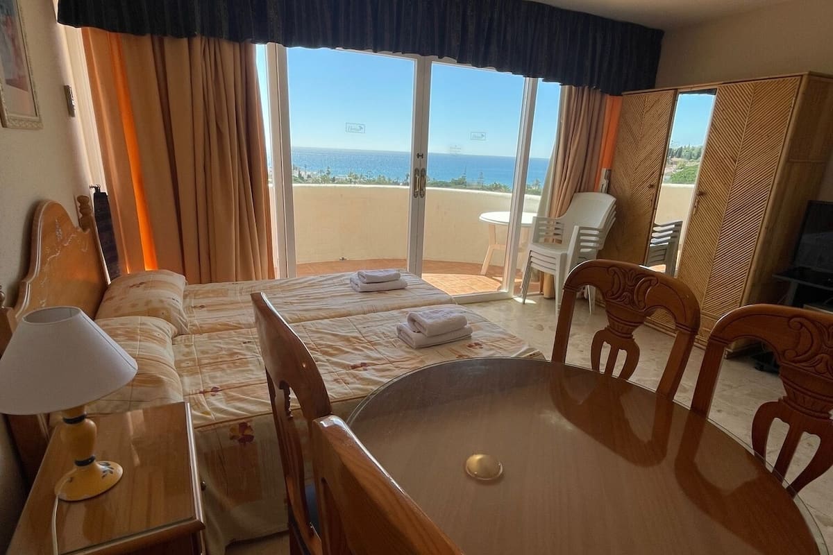 A hotel room with a bed, a dining table with chairs, a desk with a lamp, a wardrobe, and a balcony with a view of the sea.