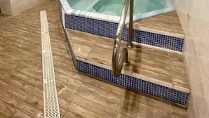 Un jacuzzi con piso de madera y paredes de azulejos, una puerta de madera y una rejilla en la pared.