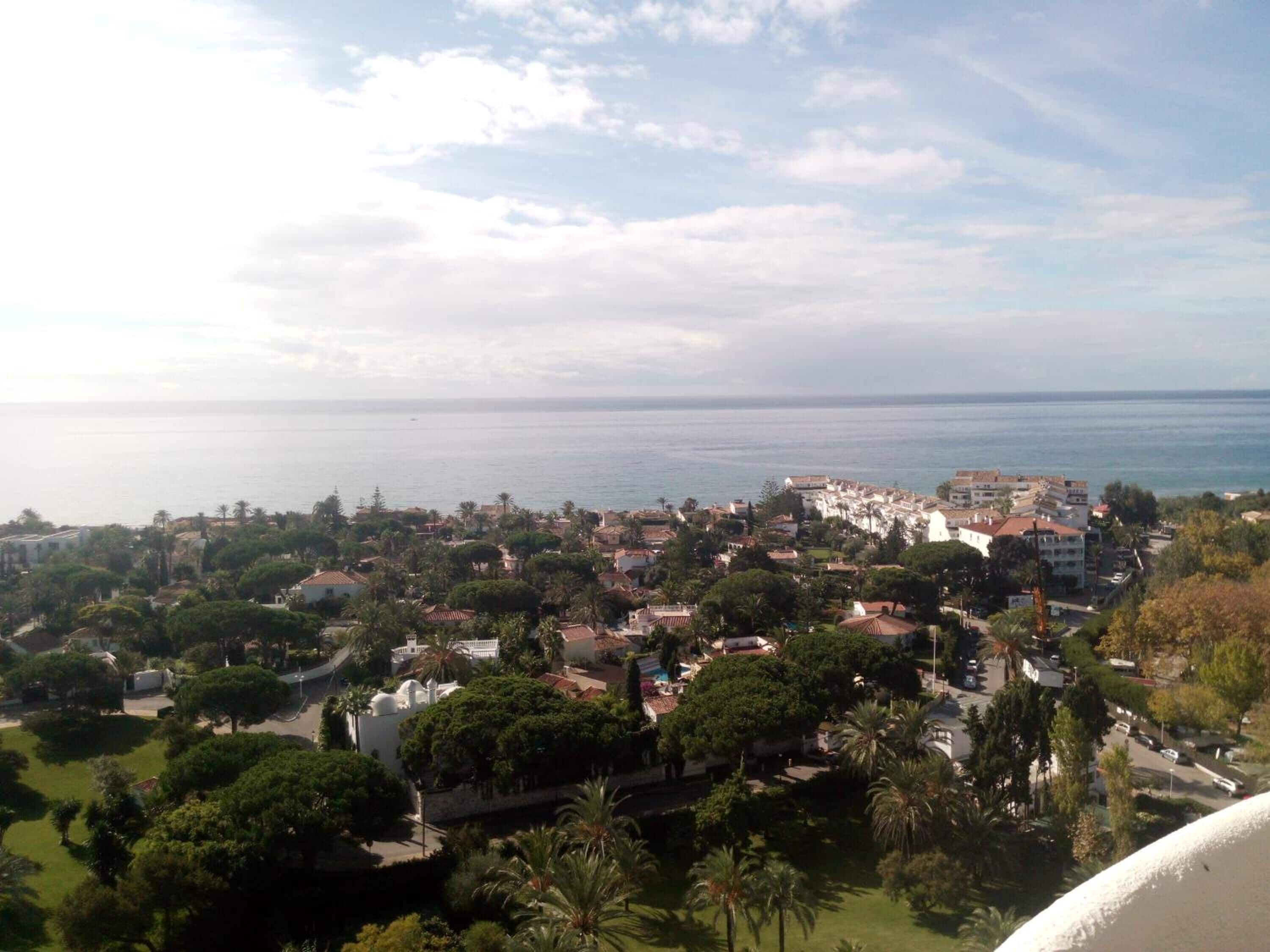 A coastal town with a mix of residential buildings and greenery, a clear view of the sea, and a prominent white building with a dome.