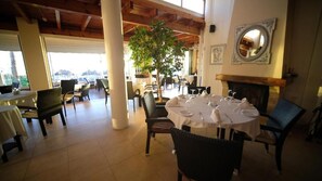 Un área de comedor interior con mesas redondas dispuestas para los comensales, una chimenea y un ventanal con vistas al exterior.