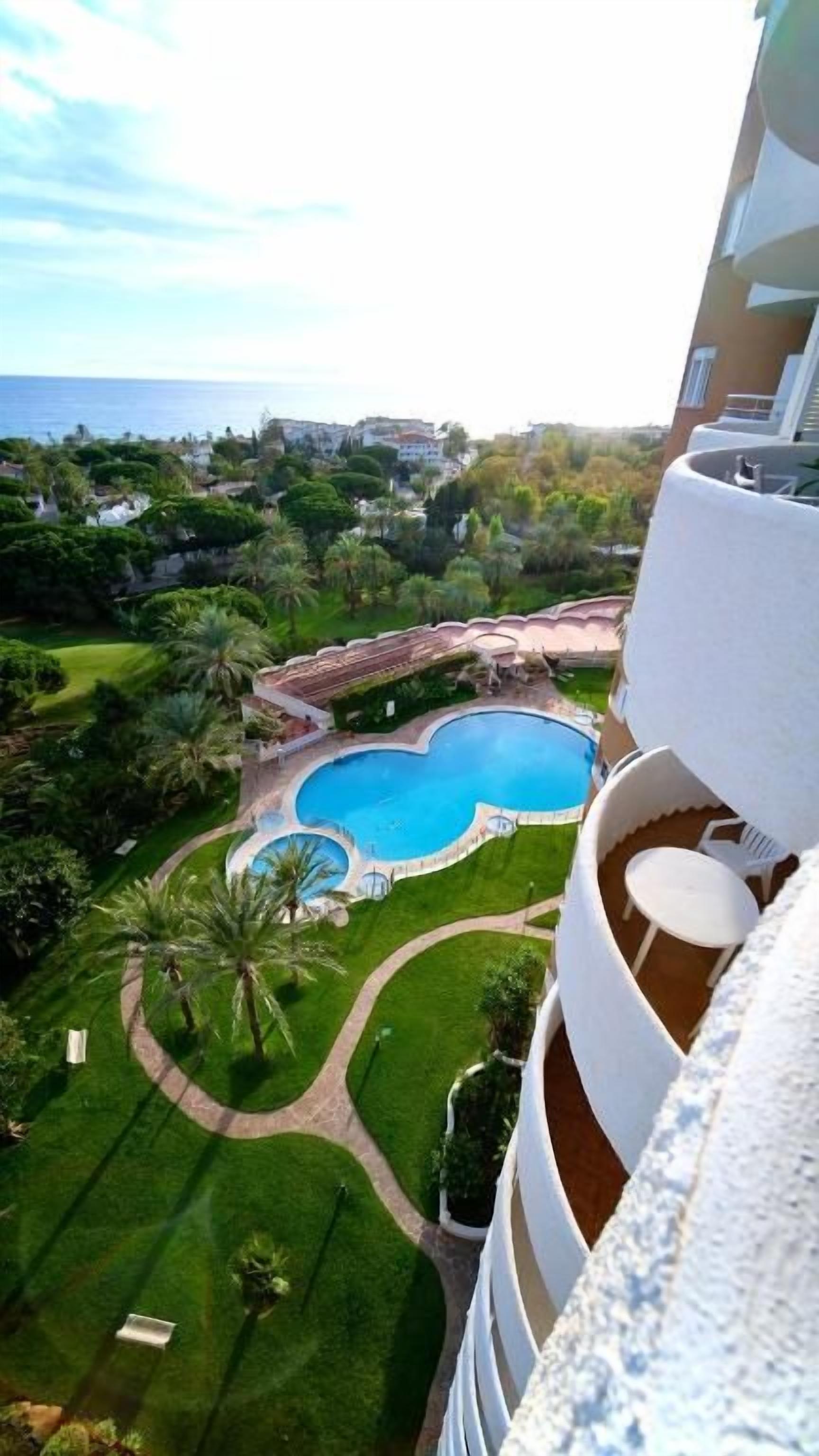 Une piscine entourée d’une pelouse bien entretenue, avec vue sur la mer et une zone résidentielle en arrière-plan.
