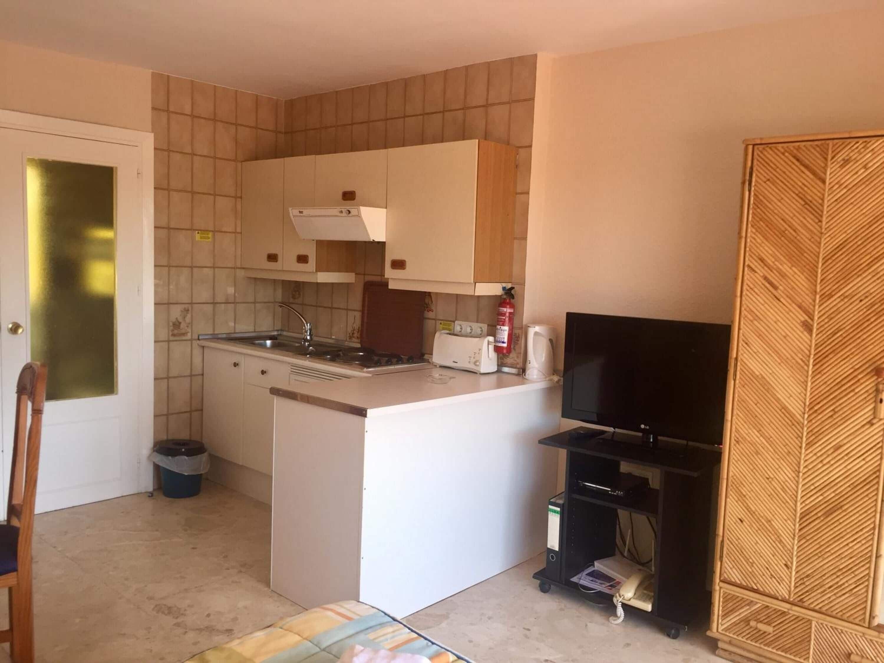 A compact kitchen with a central island, a built-in stove, a sink, and a toaster. There is a black TV on a stand to the right and a wooden cabinet with a leaf pattern to the right of the kitchen.