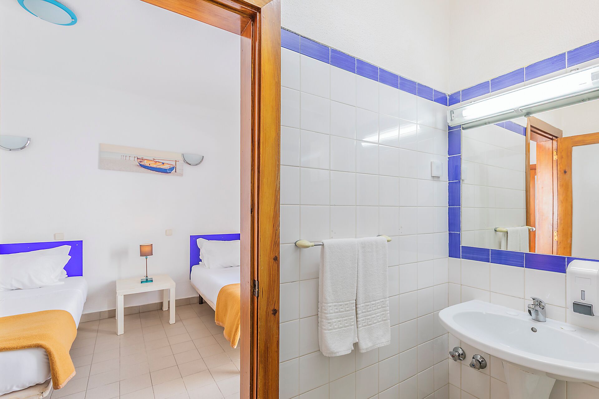 Quarto de hotel com duas camas, uma pequena mesa de cabeceira com abajur, um banheiro com pia e espelho, e um porta-toalhas fixado na parede.