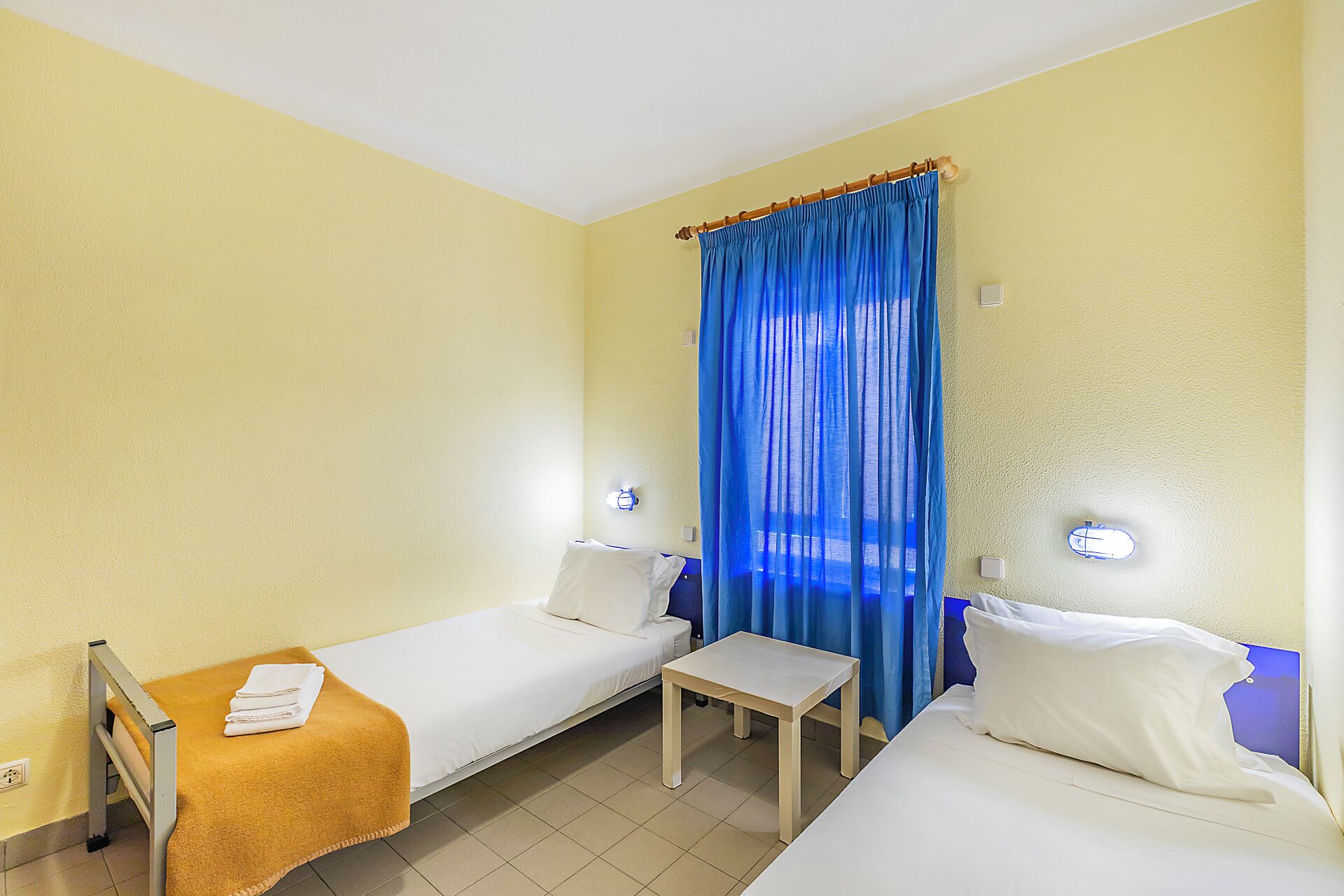 A hotel room with two single beds, a small wooden table, and a window with blue curtains.