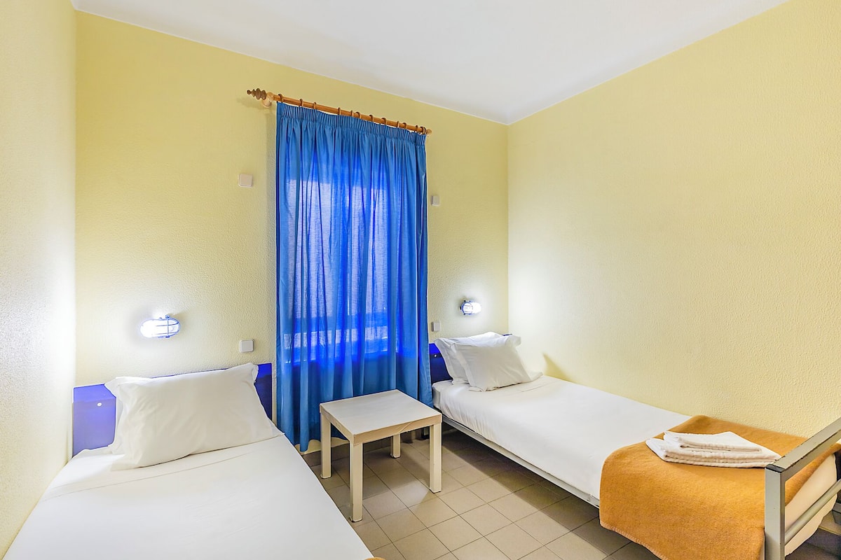 Een kleine kamer met twee eenpersoonsbedden, een tafeltje en een blauw gordijn.