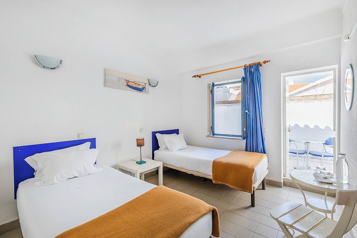 Een hotelkamer met twee bedden, een raam met gordijnen, een klein tafeltje met een stoel en uitzicht naar buiten.