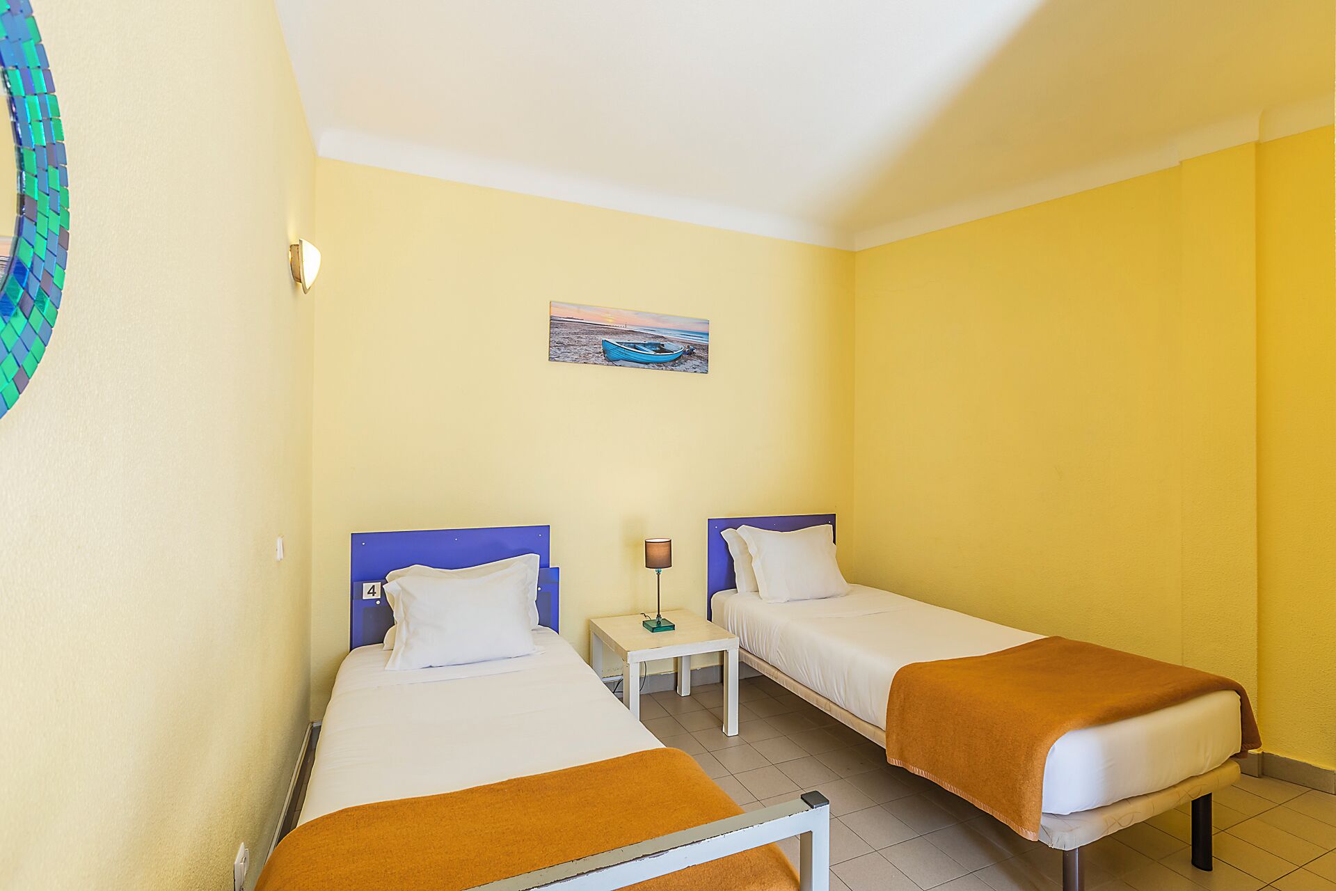 Una camera d'albergo con due letti singoli, un comodino con una lampada, un quadro appeso al muro e un pavimento piastrellato.
