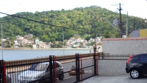A parking area with a fence, a car, and a view of a lake and houses in the background. - Kitnet em São Vicente (Sao Vicente)
