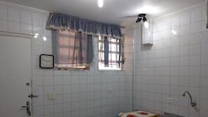 A small kitchen with white tiled walls, a window with a checkered curtain, a clock, and a sink. - Kitnet em São Vicente (Sao Vicente)