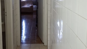 A narrow hallway with white tiled walls and a tiled floor leading to a bedroom with a bed and a window with curtains. - Kitnet em São Vicente (Sao Vicente)