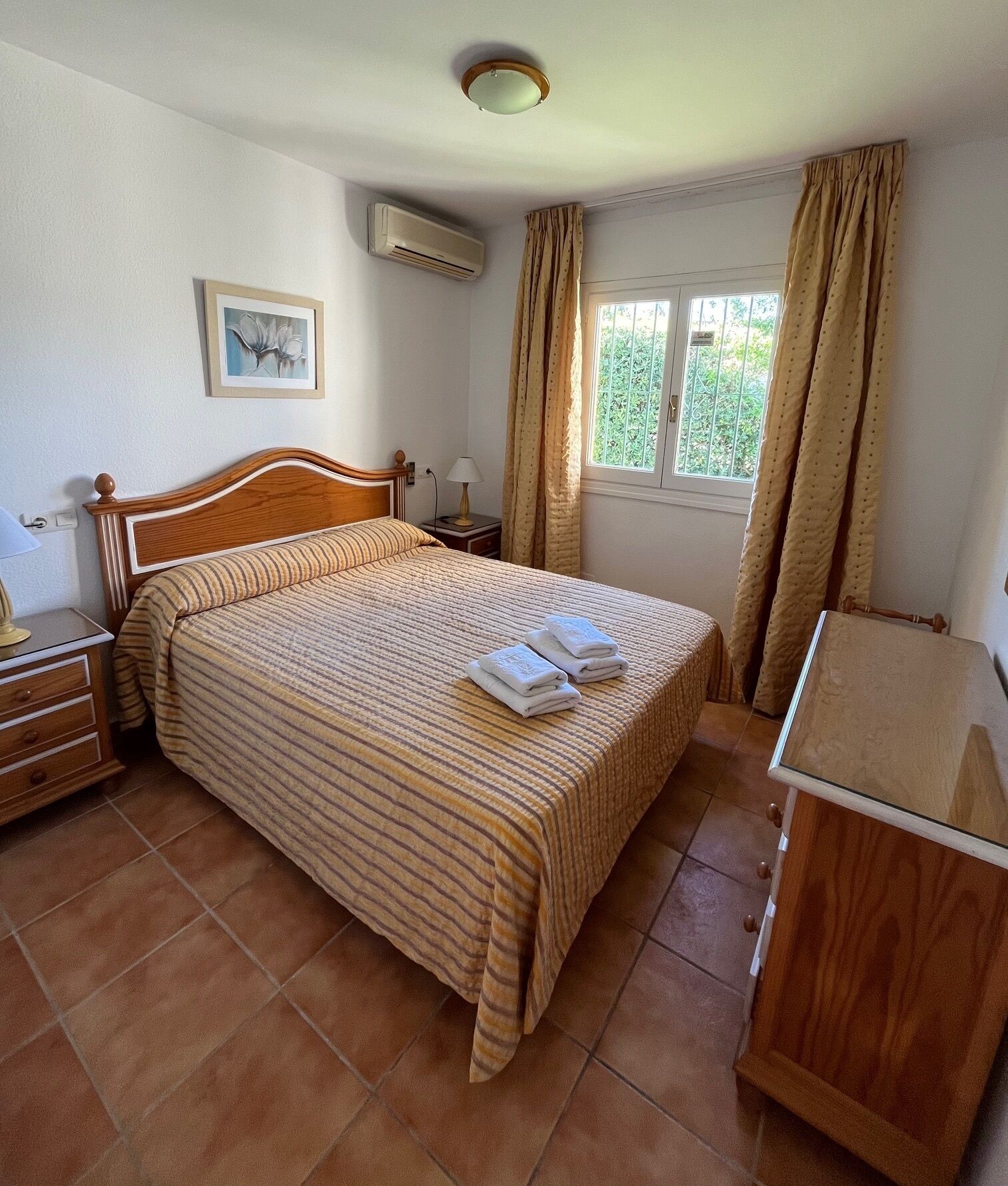 A hotel room with a large bed, a nightstand, a lamp, an air conditioning unit, a window with curtains, and a painting on the wall.